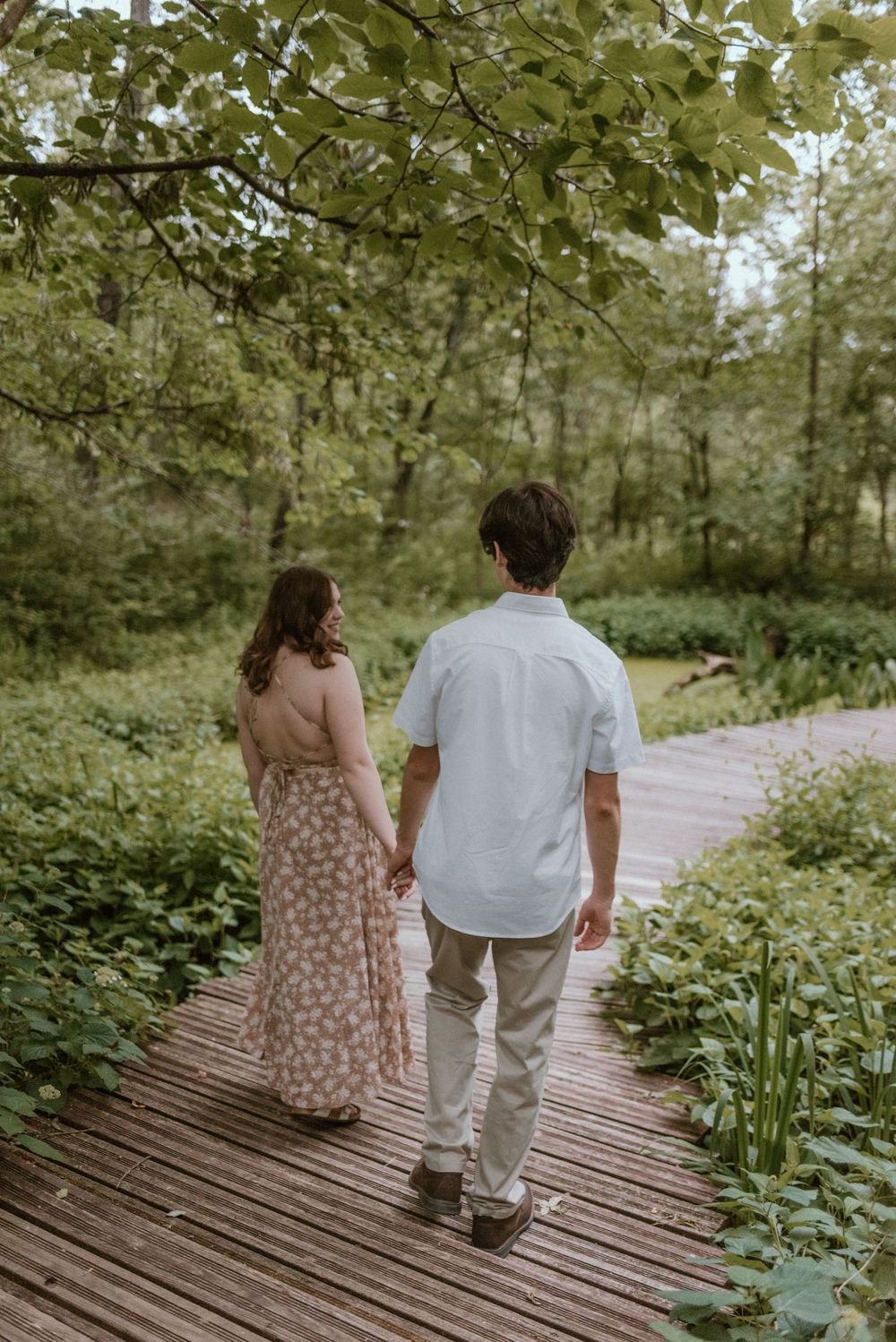 Two people walk together on a wooden boardwalk surrounded by lush greenery.