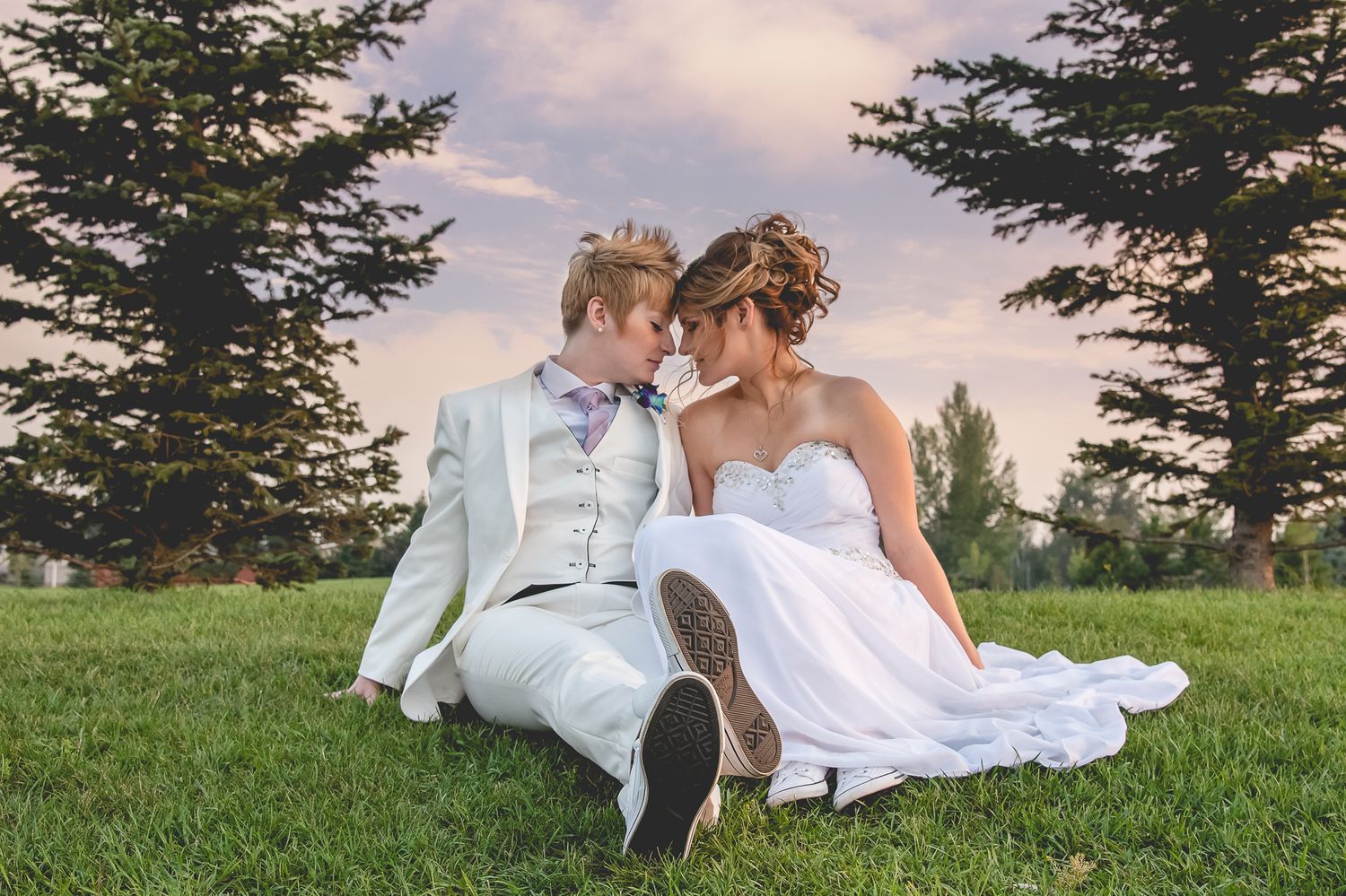 A couple in wedding attire sits together on lush grass with evergreen trees in the background.