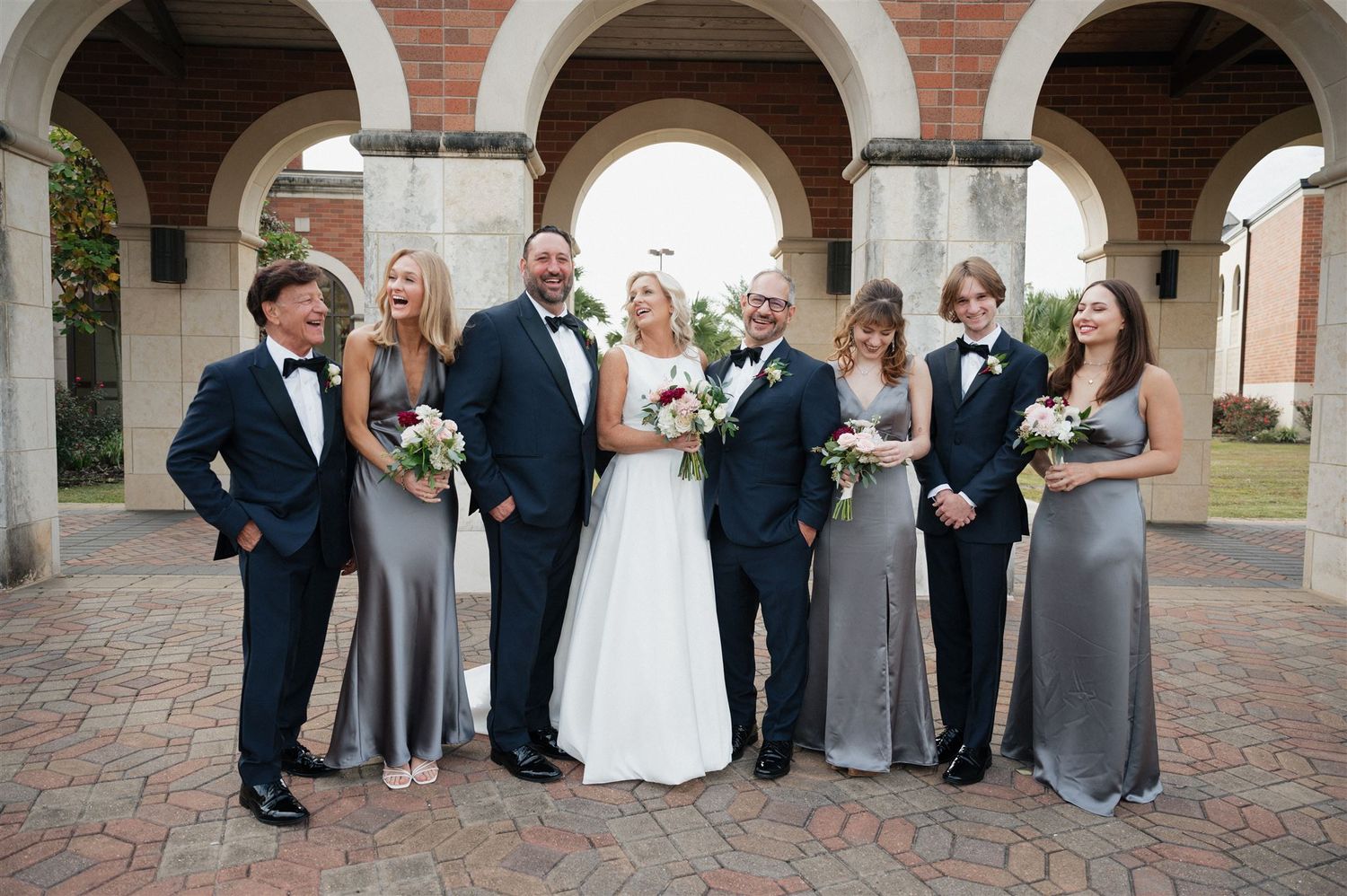 Wedding party poses together under arched columns wearing formal attire in grey and black.