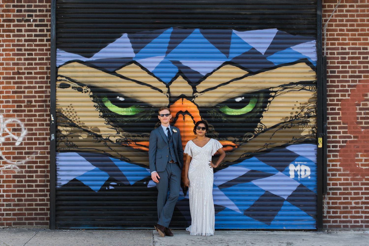 Two people pose in front of a large street art mural featuring geometric owl eyes on a brick wall.