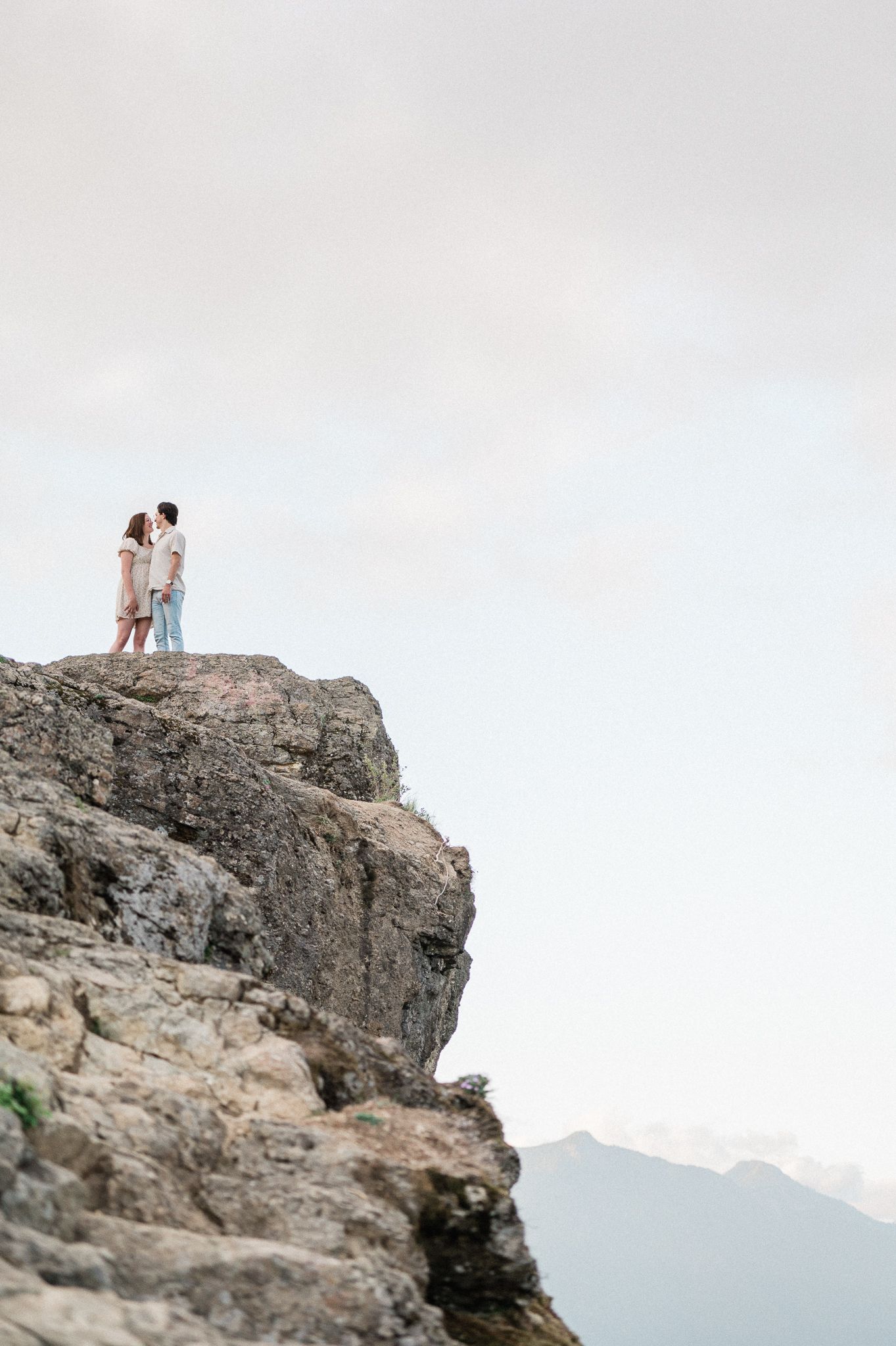 Couple stand atop a dramatic rocky cliff overlooking mountain vistas during a sunset.