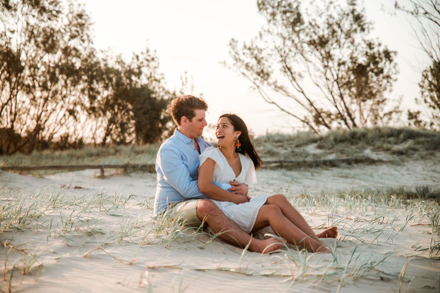 Happy couple sitting together on sandy dunes during sunset photoshoot.