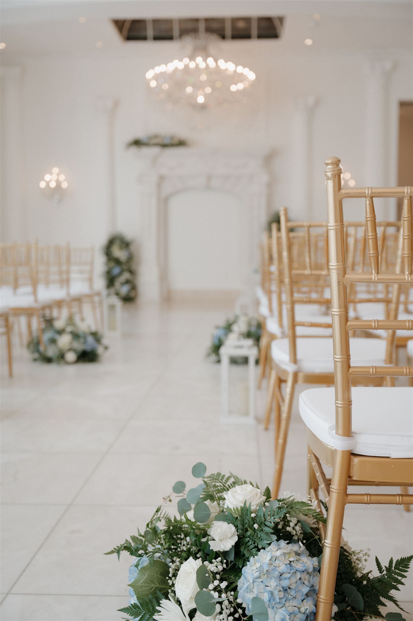 Elegant wedding venue with gold chiavari chairs, crystal chandeliers, and blue and white floral arrangements along the aisle.