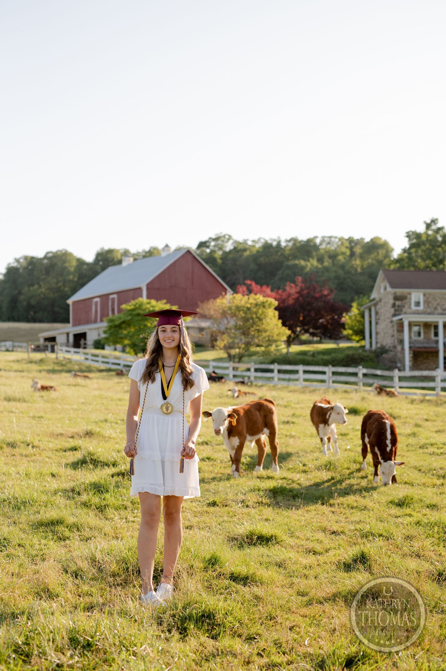Graduate poses in front of red barn and farmhouse with cattle grazing in sunlit field.