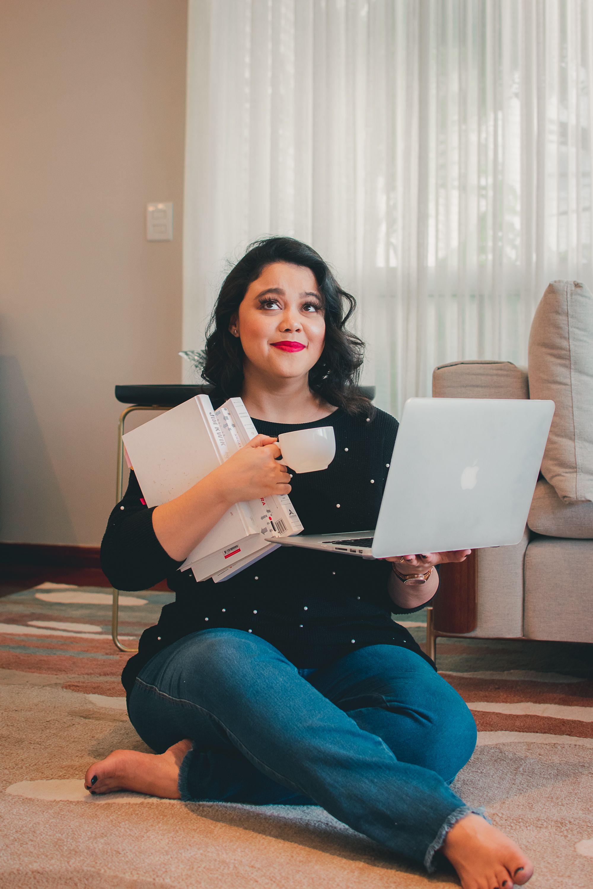 Someone sits cross-legged on floor with laptop and coffee in casual home setting.
