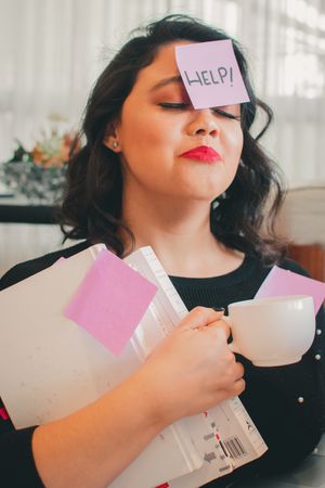 Someone with red lipstick has HELP sticky note on forehead while holding papers and coffee.