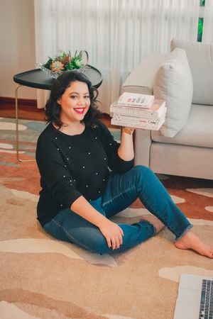 Person sitting on carpet holding books while wearing black sweater and jeans in cozy home setting.