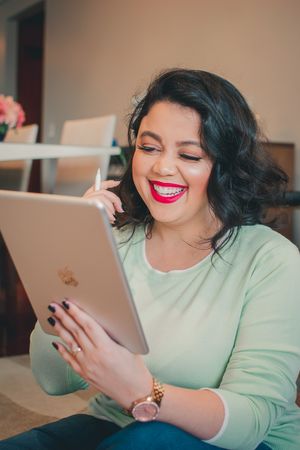 Woman in mint sweater smiling while looking at tablet device in living room setting.