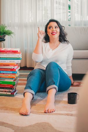 Reader sitting on floor with large stack of books raising finger in thoughtful gesture.