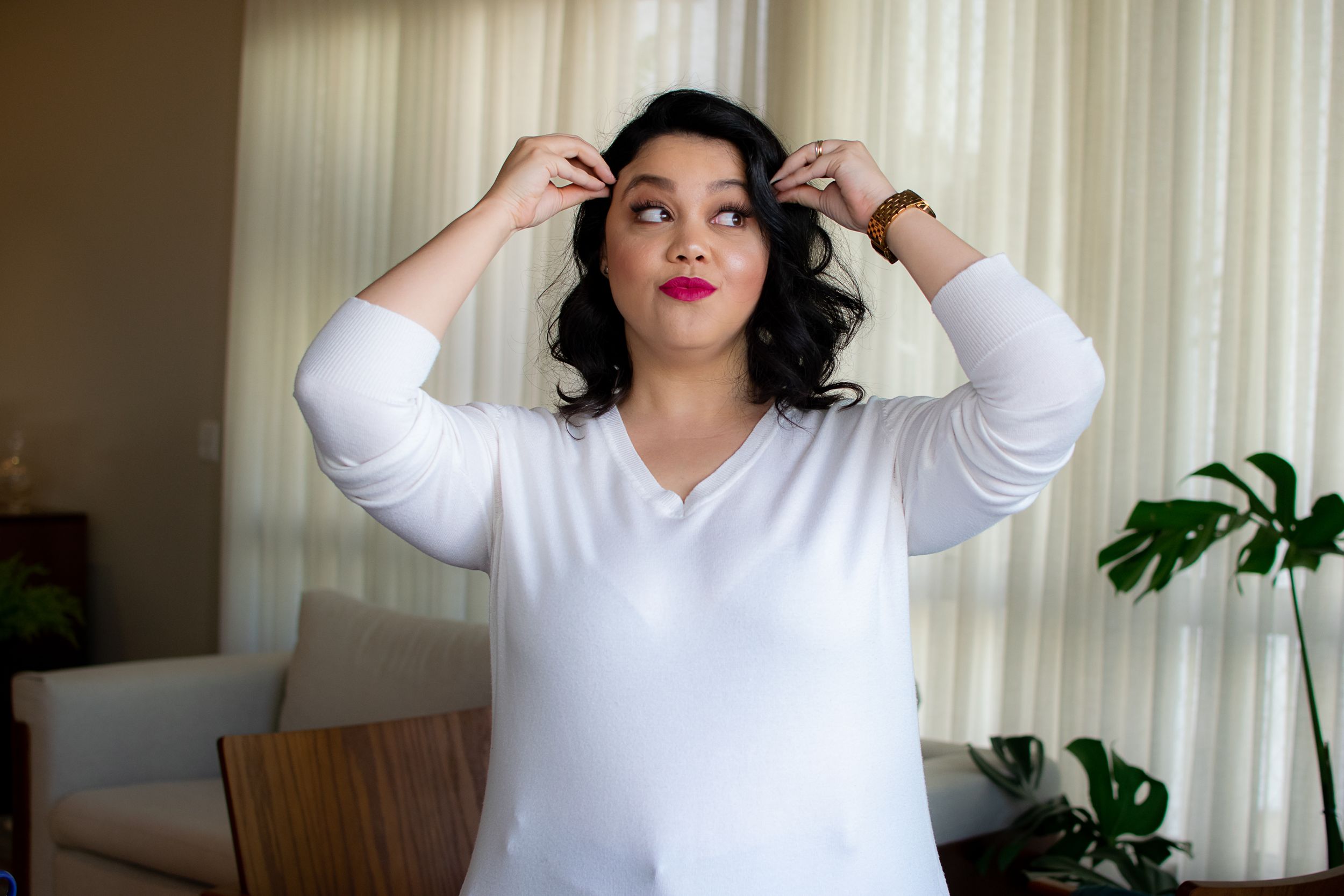 Person in white top making playful expression and gestures against curtained backdrop.