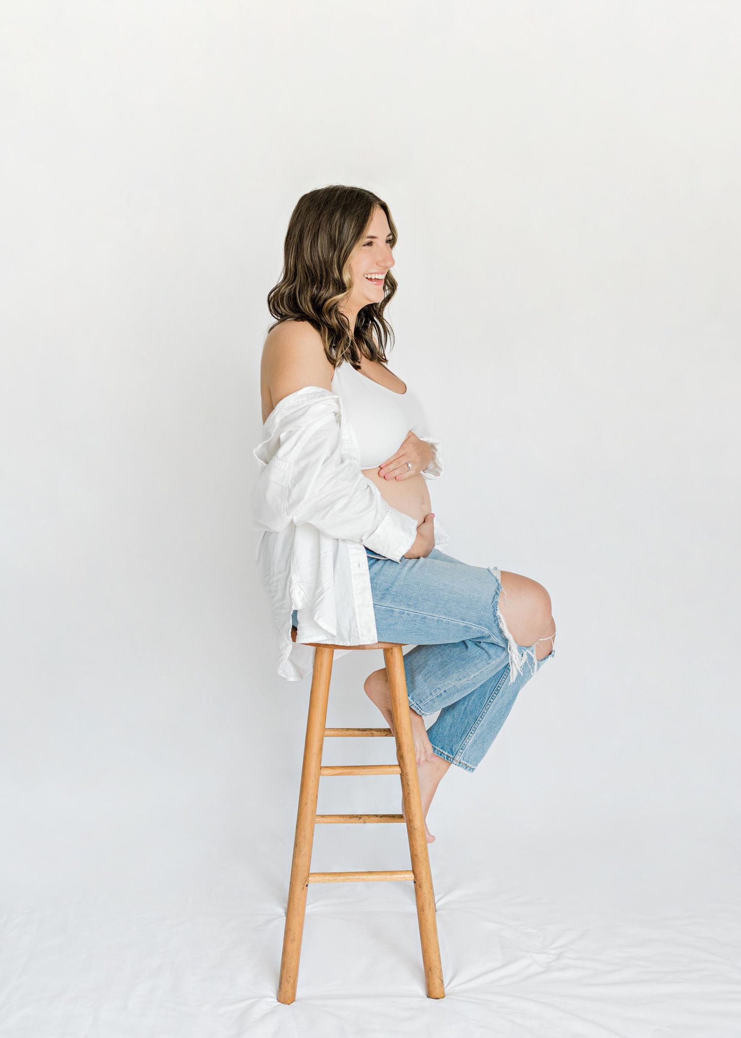 Maternity photo of a woman in white top and ripped jeans sitting on wooden stool against white background.