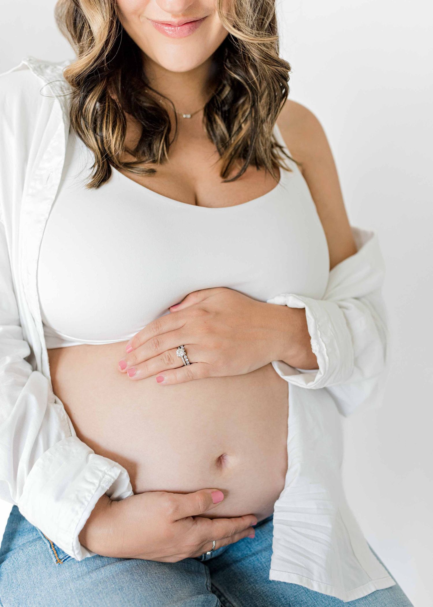 Close up maternity photo showing pregnant belly with hands and white top against neutral background.