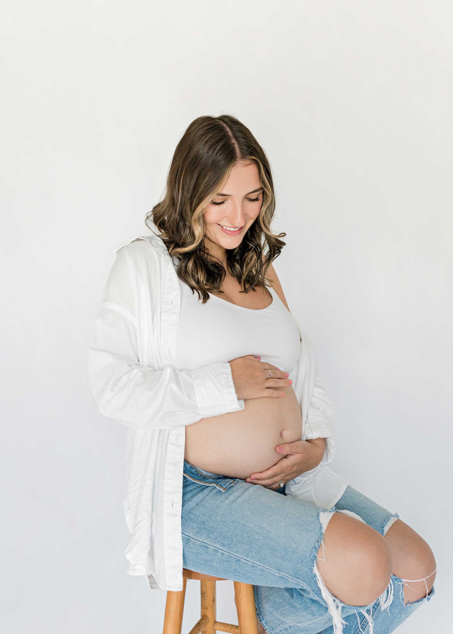 Natural maternity portrait with woman in white cardigan and ripped jeans on wooden stool.