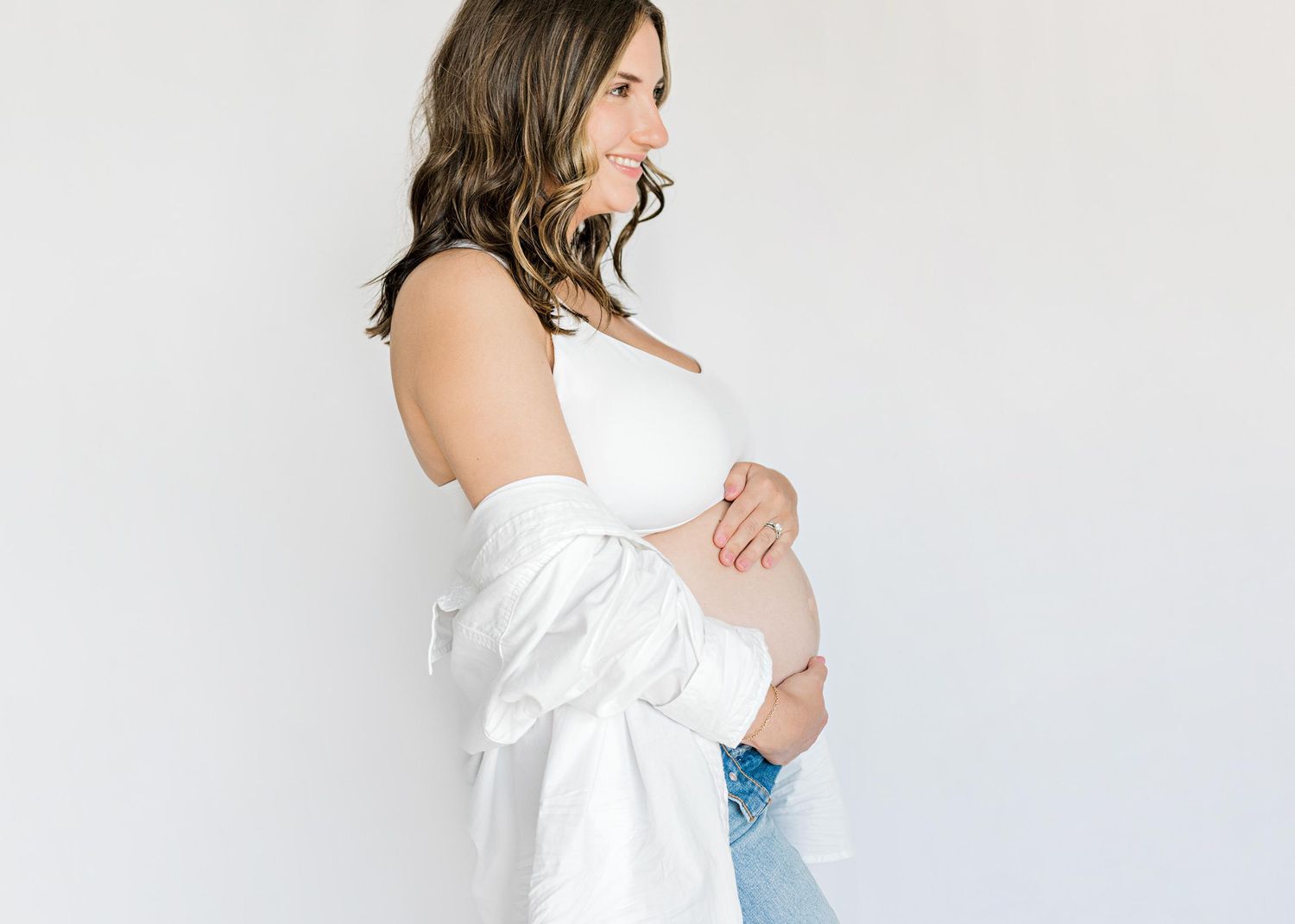 Artistic maternity pose featuring white shirt and denim against minimalist background.