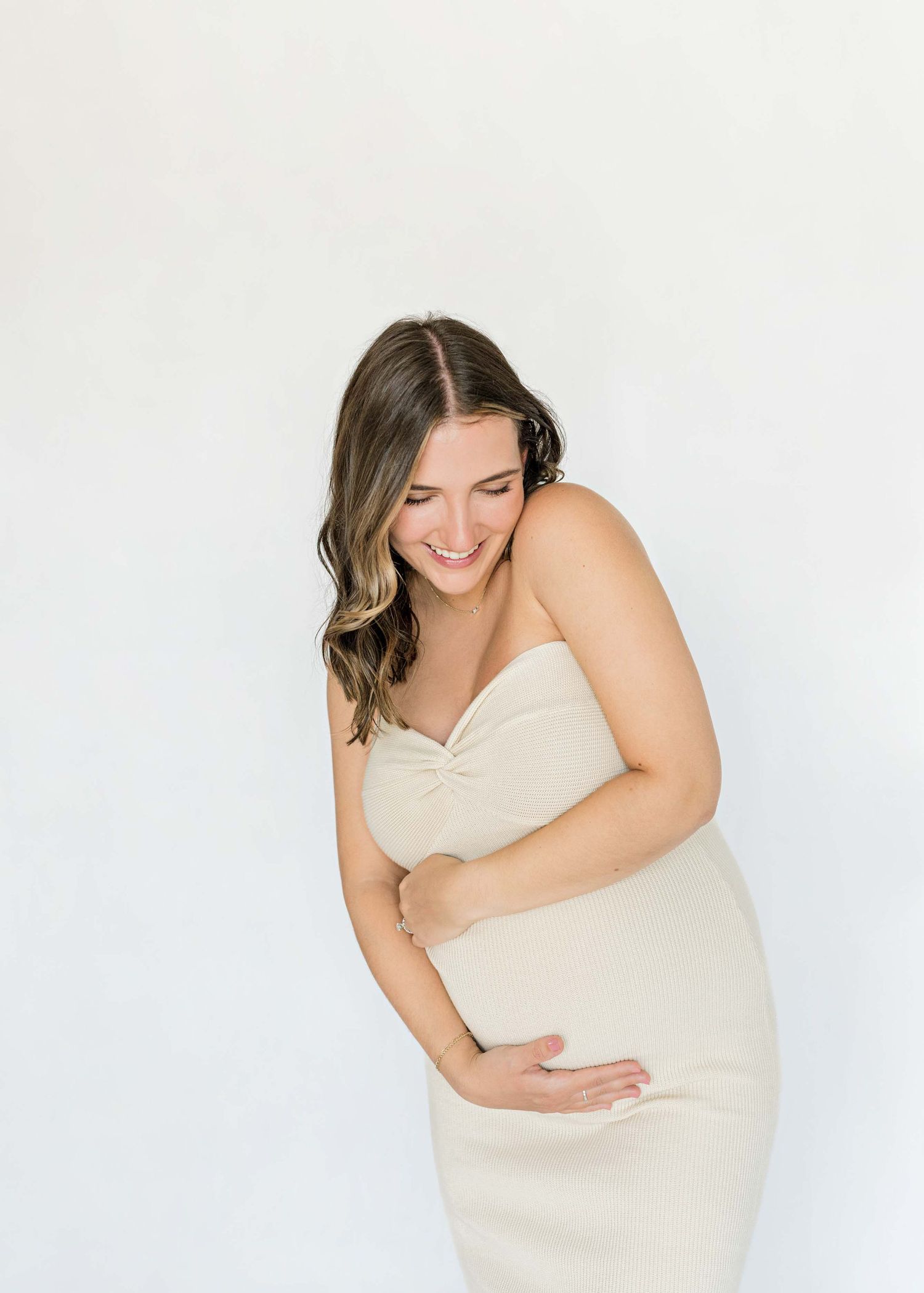 Intimate maternity portrait showing joyful expression in cream fitted dress against white backdrop.