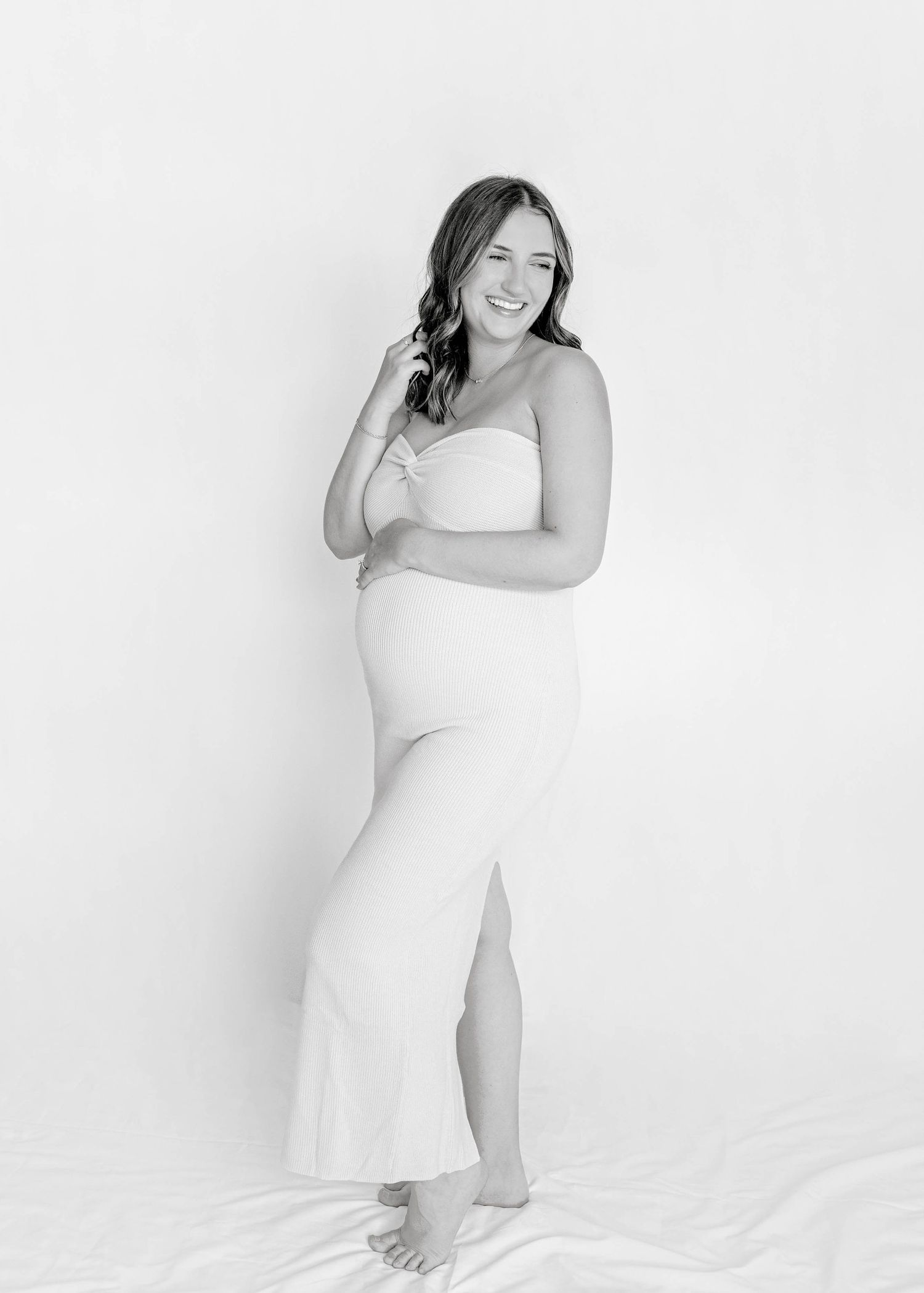 Maternity photoshoot in black and white features a woman in a white dress posing against a simple backdrop.