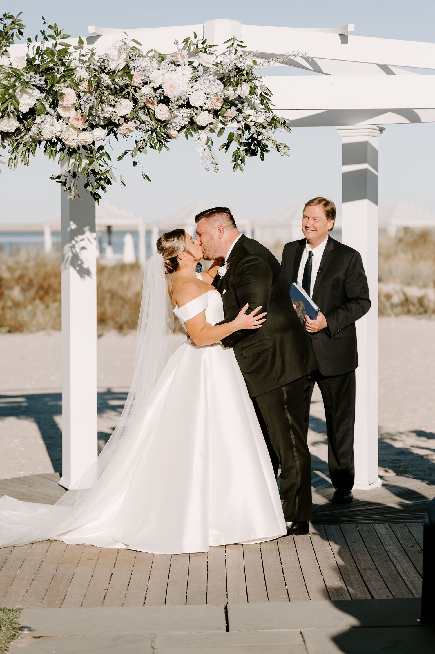 Newlyweds share a romantic moment under a white pergola on a sunny beach boardwalk.