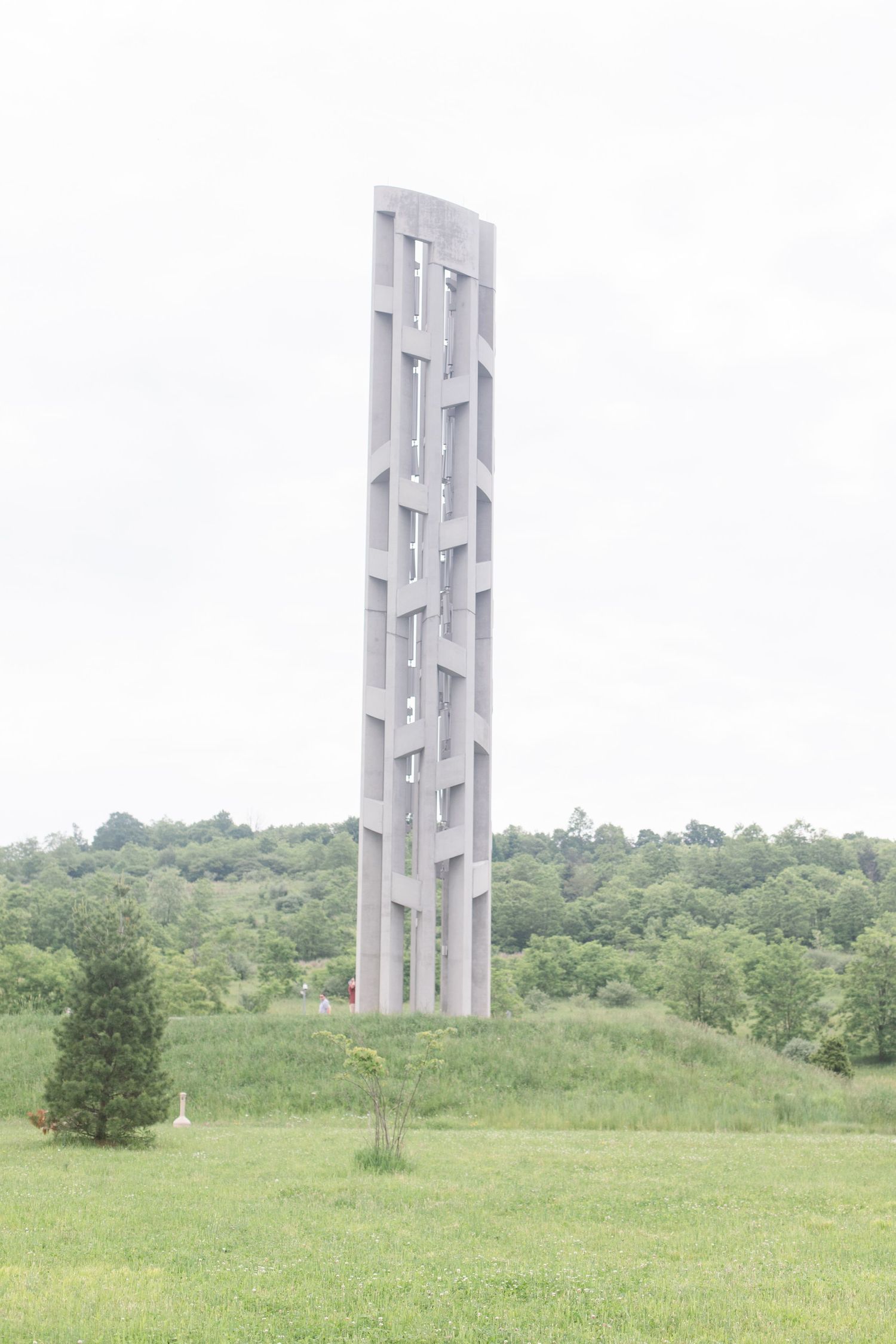 Concrete Tower of Voices memorial standing in a grassy meadow against cloudy sky.