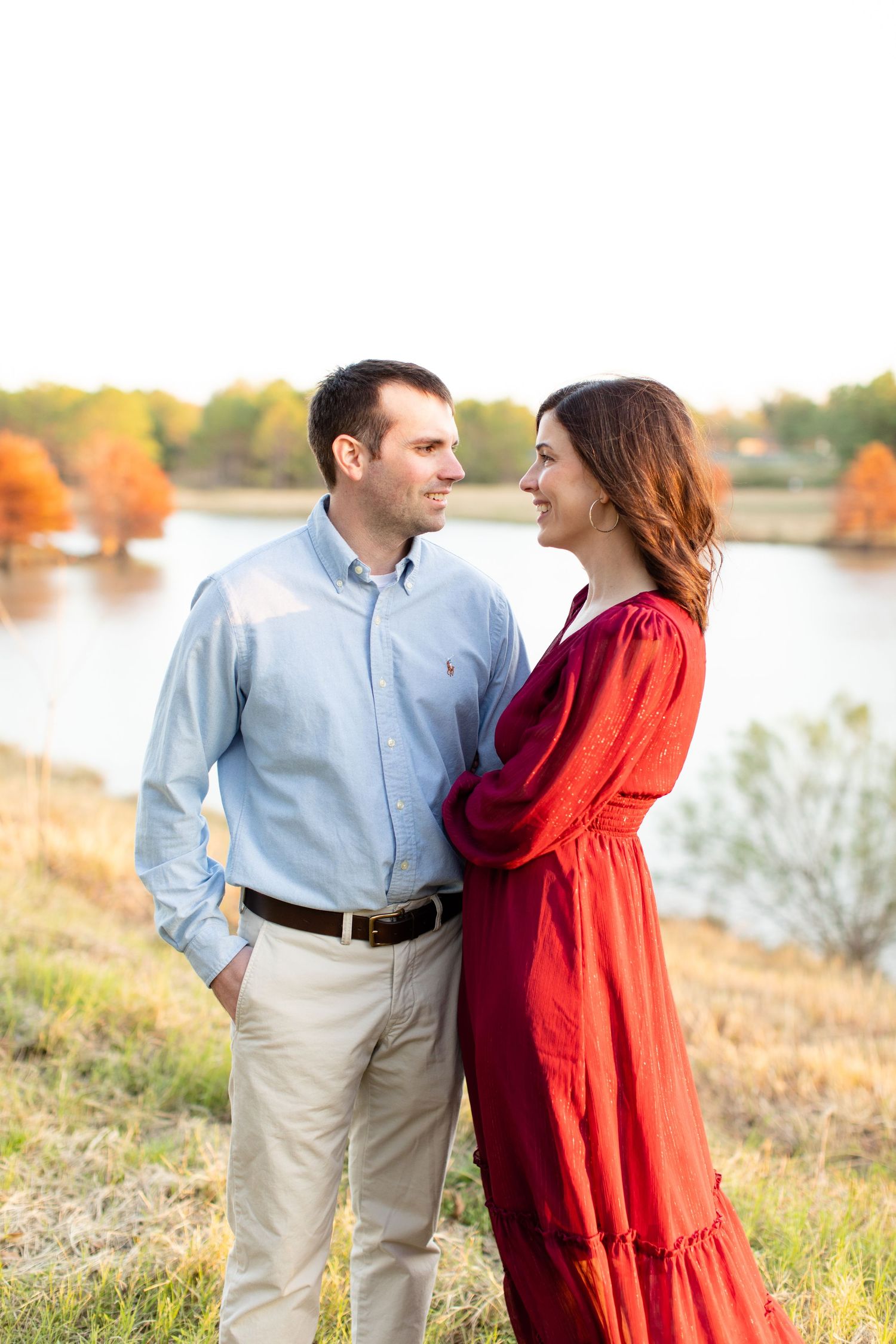 Couple in burgundy dress and blue shirt standing by lake during golden hour.