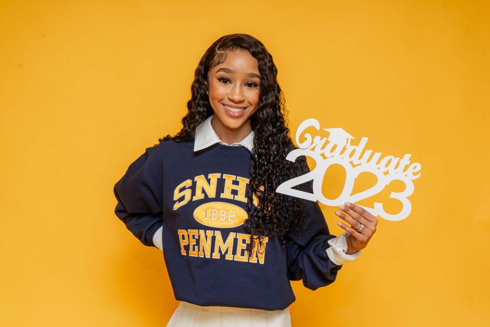 Smiling graduate in SNHU Penmen sweatshirt holds Graduate 2023 sign against vibrant yellow background.