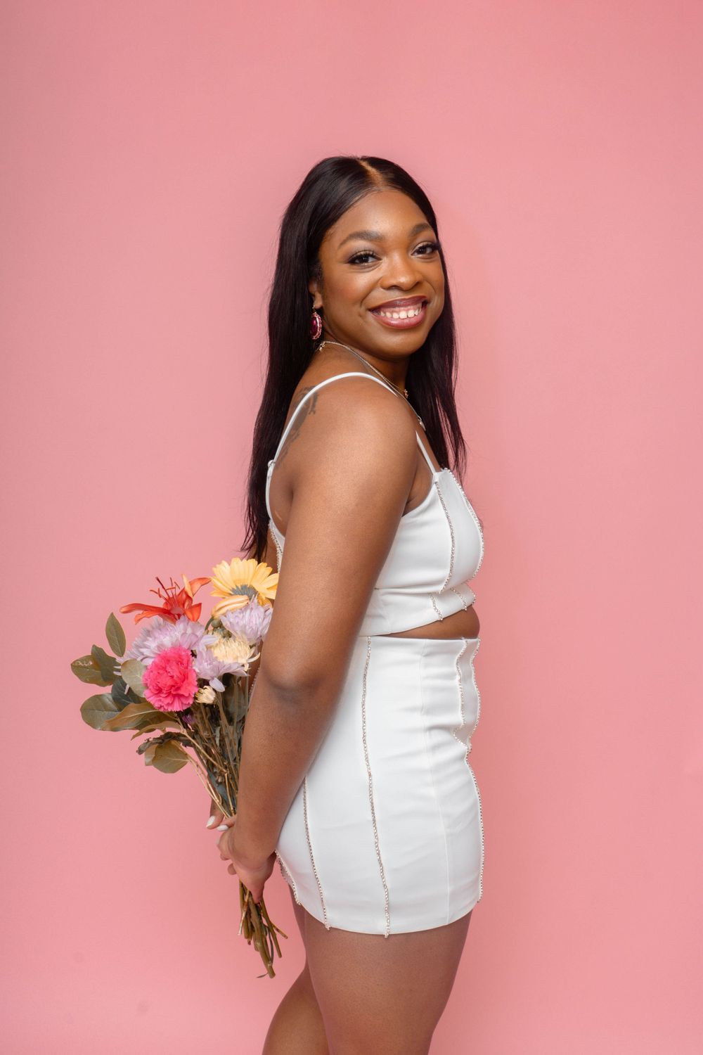 Smiling woman in white crop set holding colorful flower bouquet against pink background.