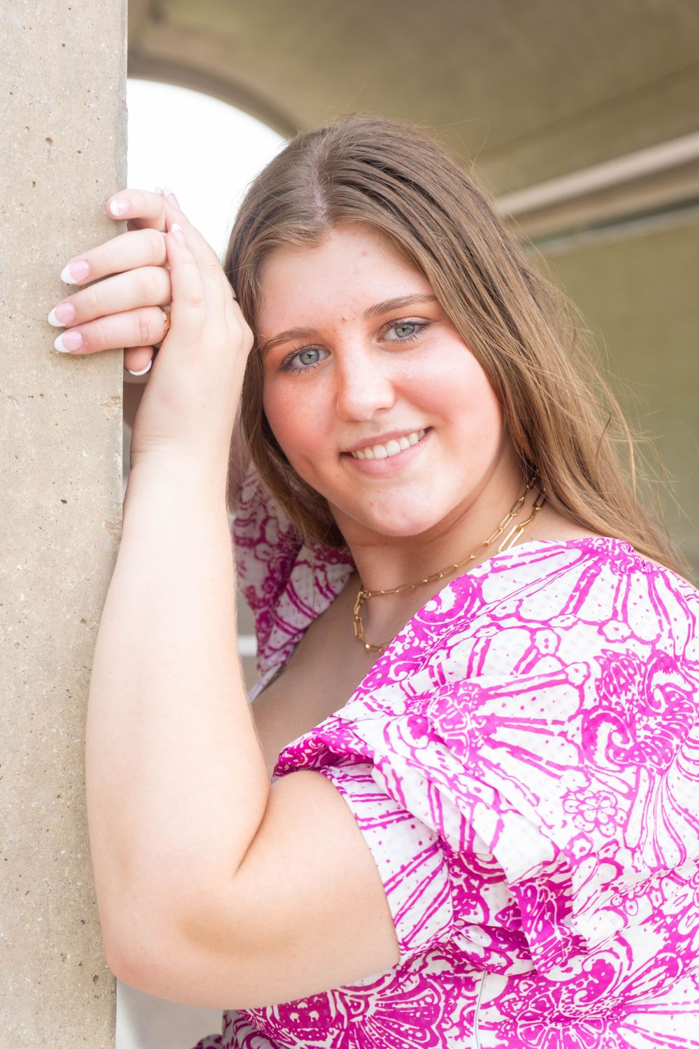 Smiling young woman with blue eyes in a pink floral top leaning against a concrete pillar outdoors.