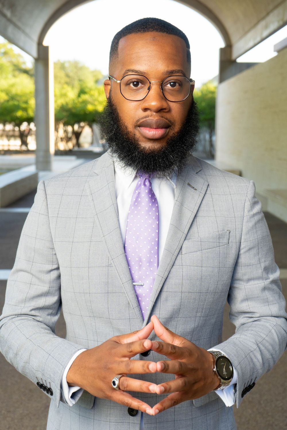 Professional Black businessman in gray suit and purple tie posing confidently under an outdoor archway.