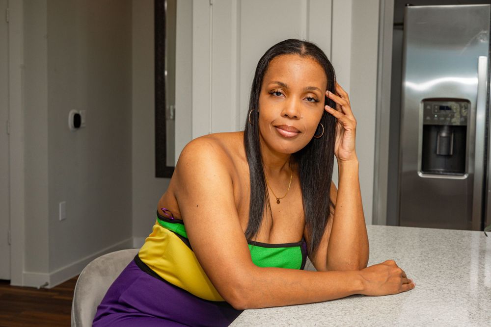 Woman in colorful green and purple top resting at kitchen counter near stainless appliances.