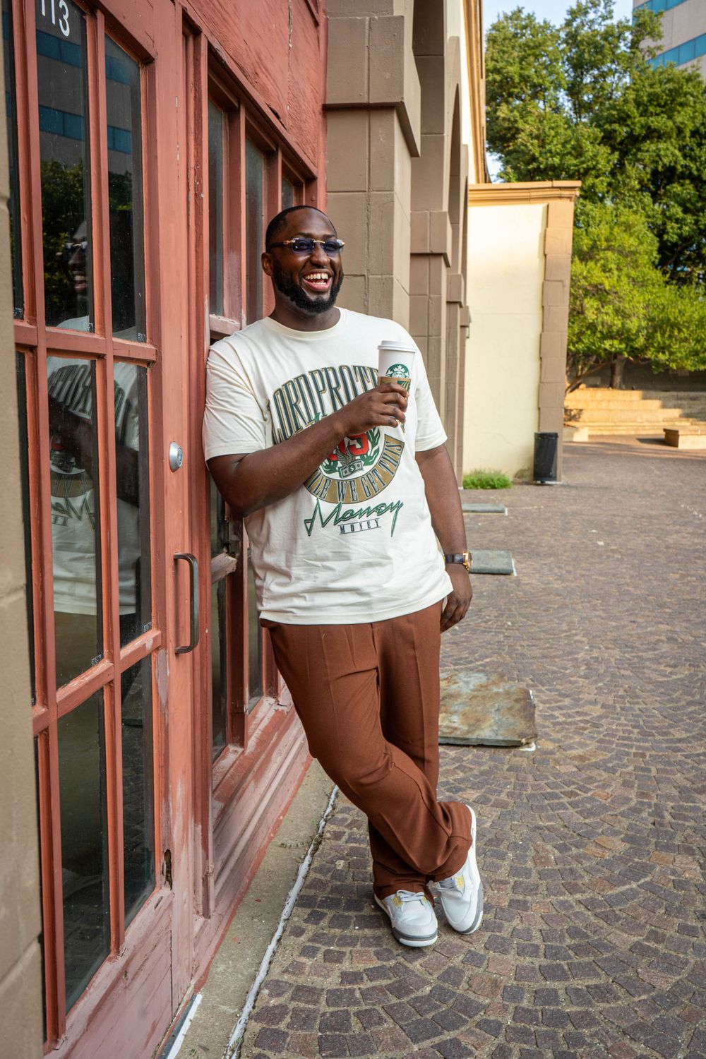 Smiling Black man in white graphic tee and brown trousers leaning against a building holding a coffee cup outdoors.