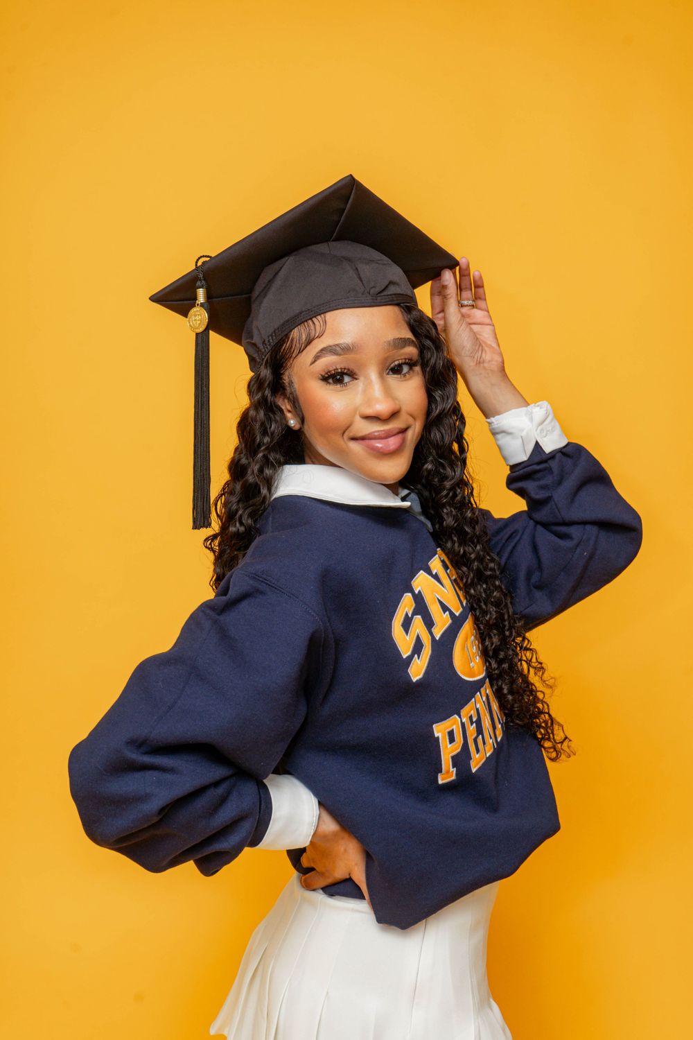 Young woman in graduation cap and navy college sweatshirt posing against bright yellow background.