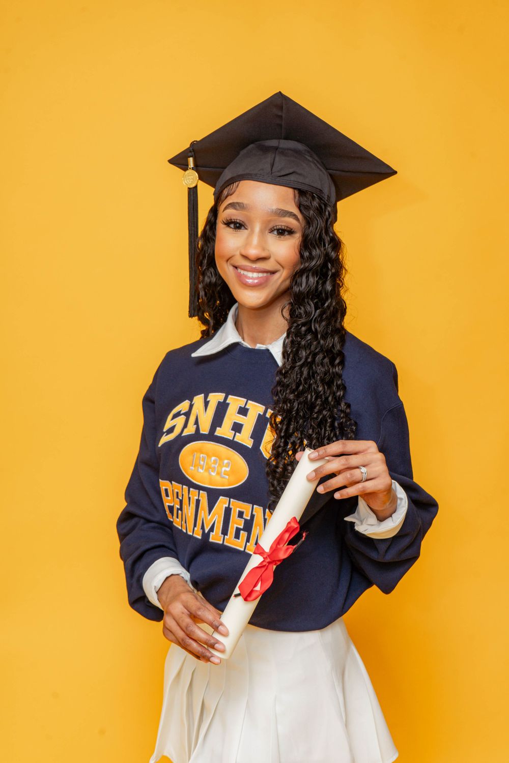 Smiling graduate in SNHU Penmen sweatshirt and cap holds diploma scroll on bright yellow background.