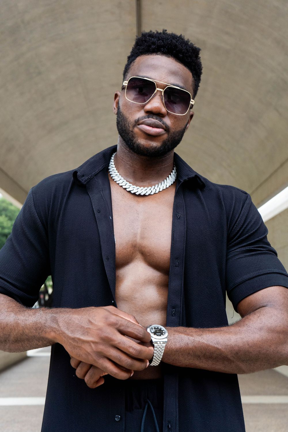 Confident Black man in open black shirt with silver chain necklace and sunglasses posing outdoors.