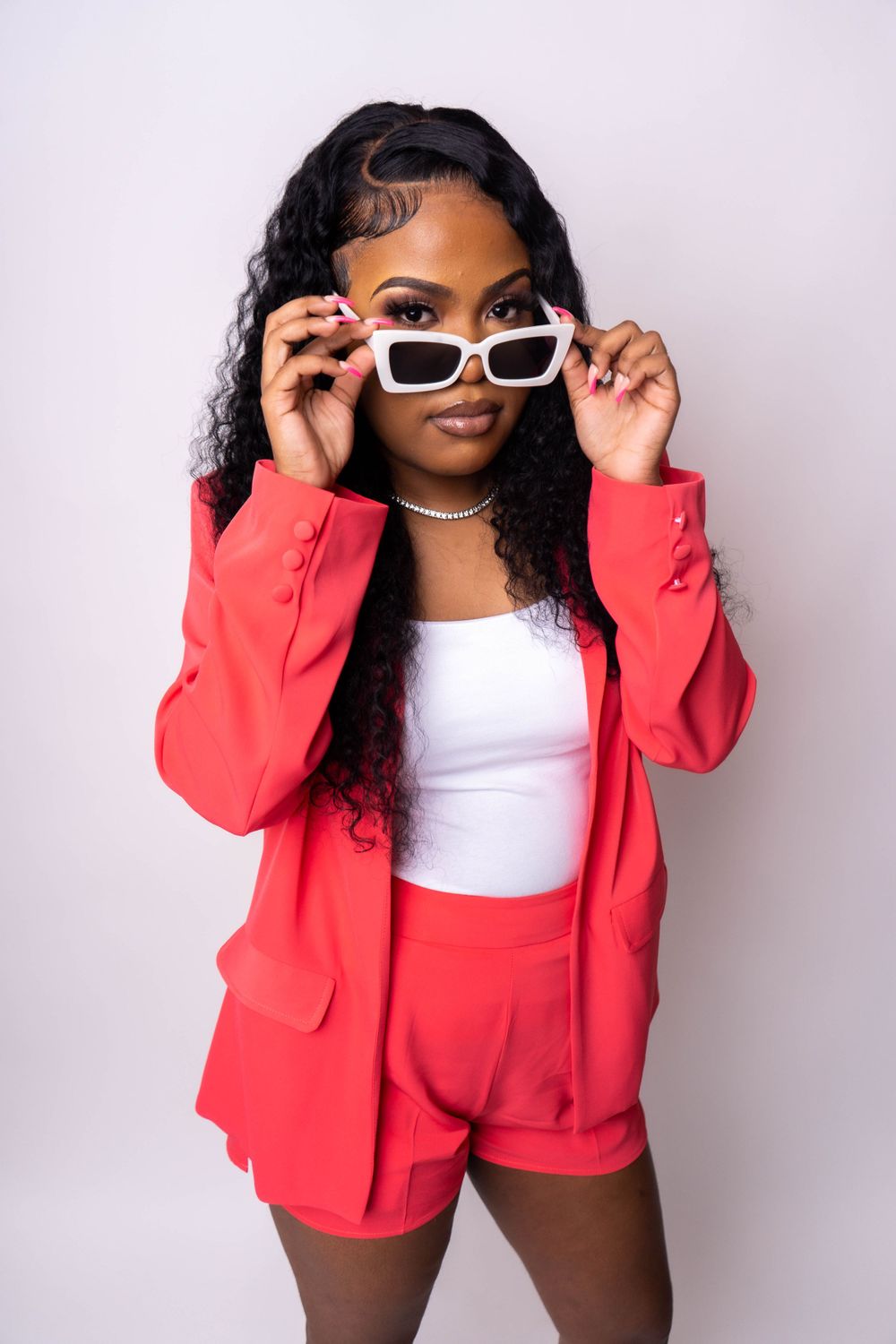 Young Black woman in a red blazer and shorts adjusting white square sunglasses against a white background.