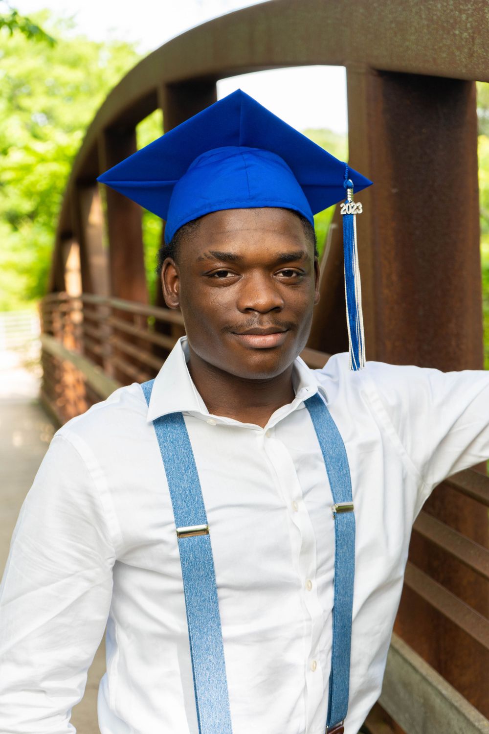 Young Black man in a blue graduation cap and white shirt with suspenders posing on a bridge.