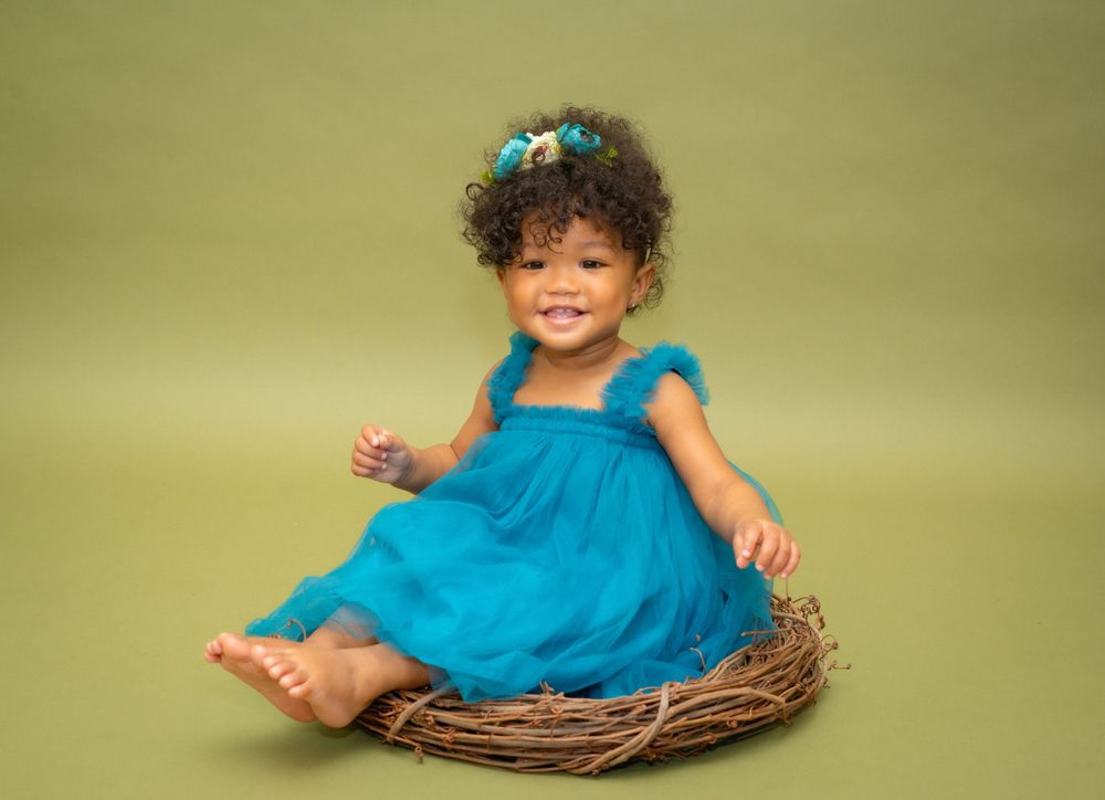 Adorable toddler girl in teal dress sitting in twig nest against green background.