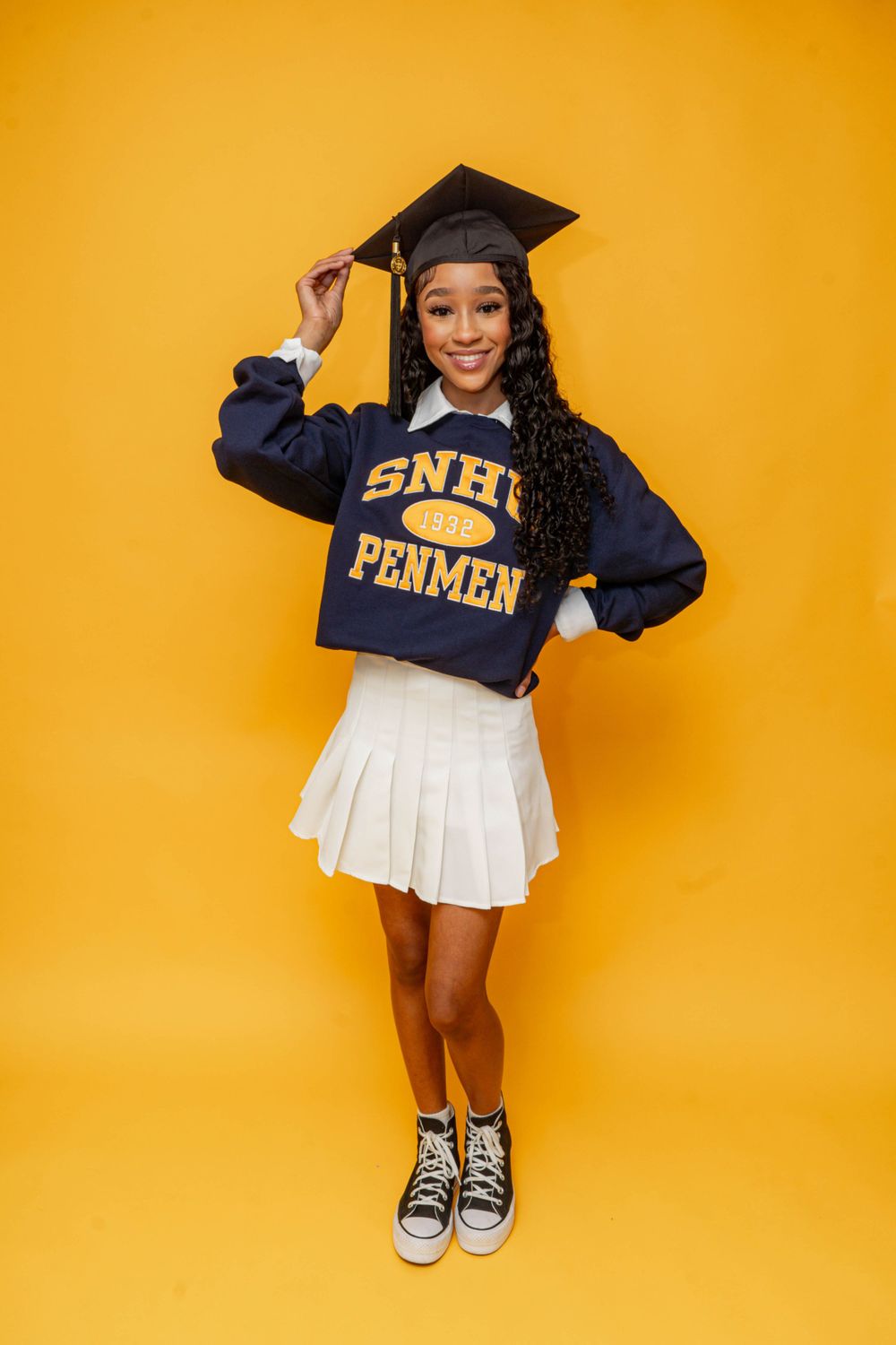 Young graduate poses in SNHU Penmen sweatshirt, white pleated skirt, and graduation cap on yellow background.