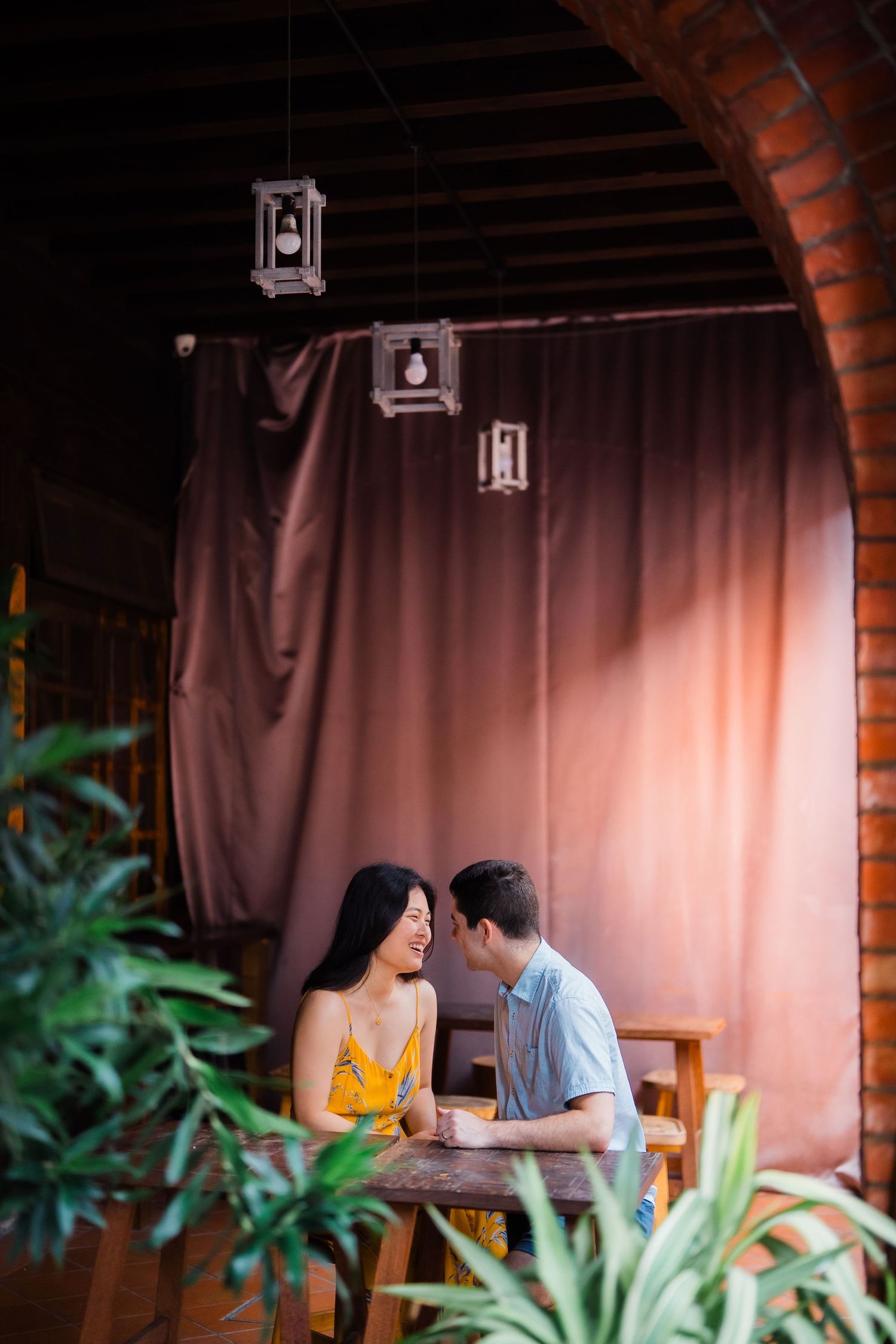Couple sharing romantic moment at cozy restaurant table with hanging lanterns and tropical plants.
