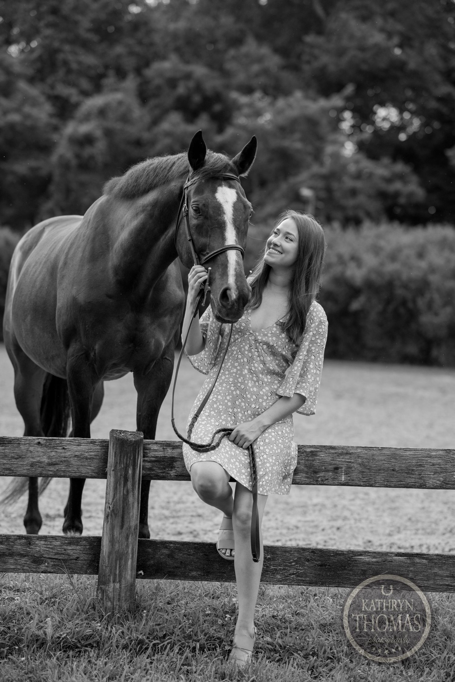 A black and white portrait of a horse and person in a dress posing by a wooden fence.