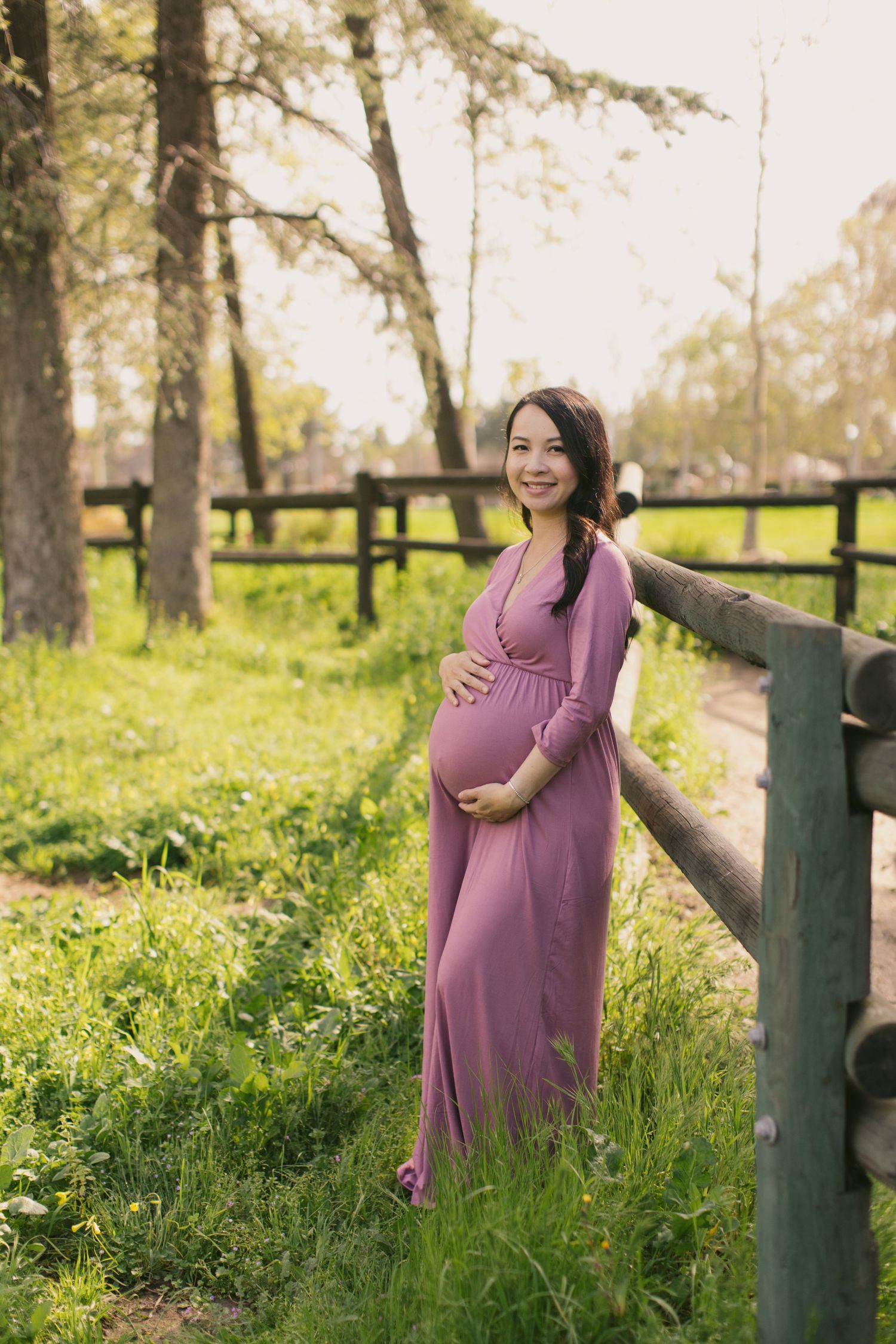 Expectant mother in flowing purple dress poses by rustic fence in sunlit forest setting.