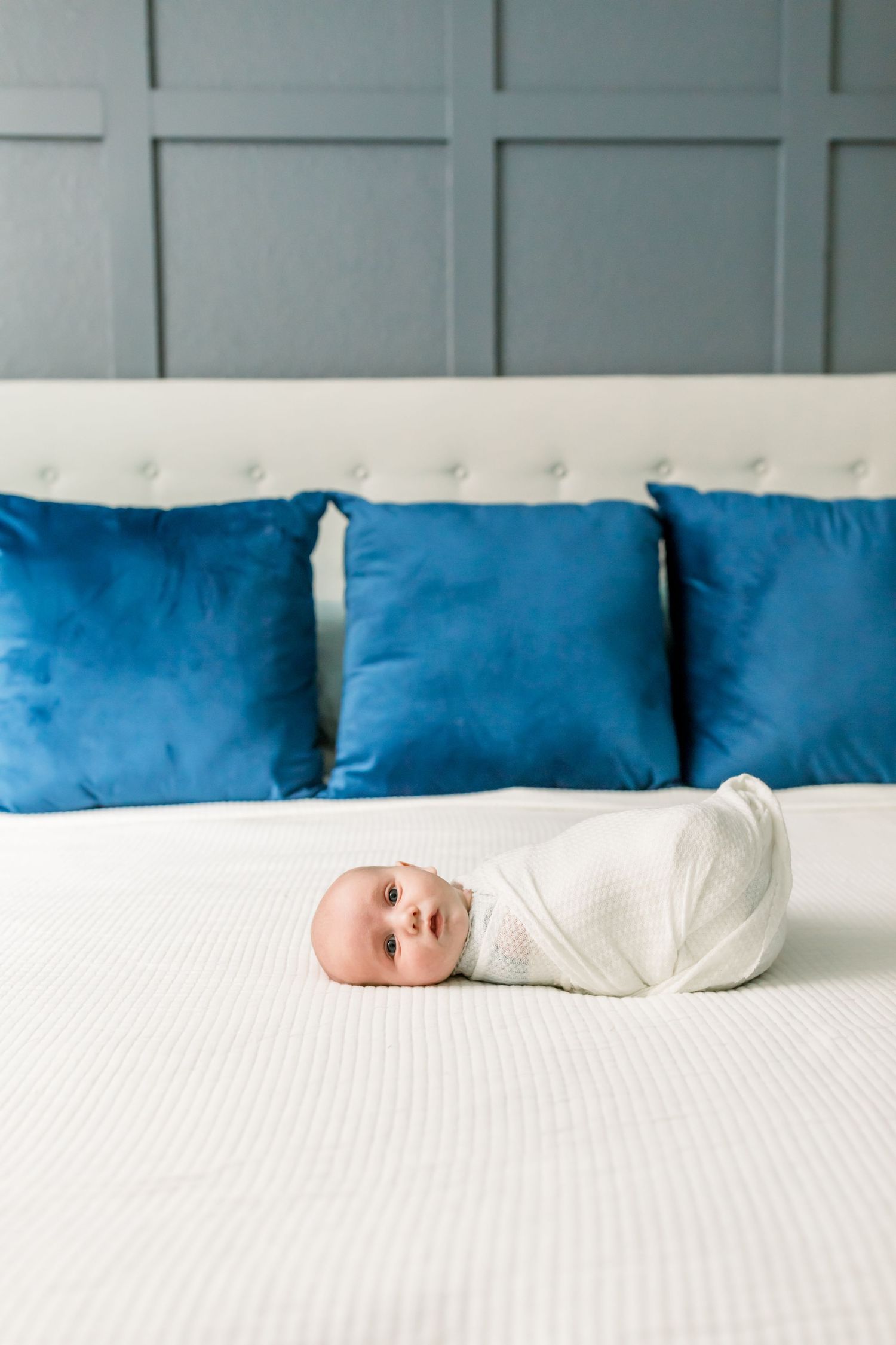 A swaddled infant rests on a white bed with blue throw pillows against a grey wall.