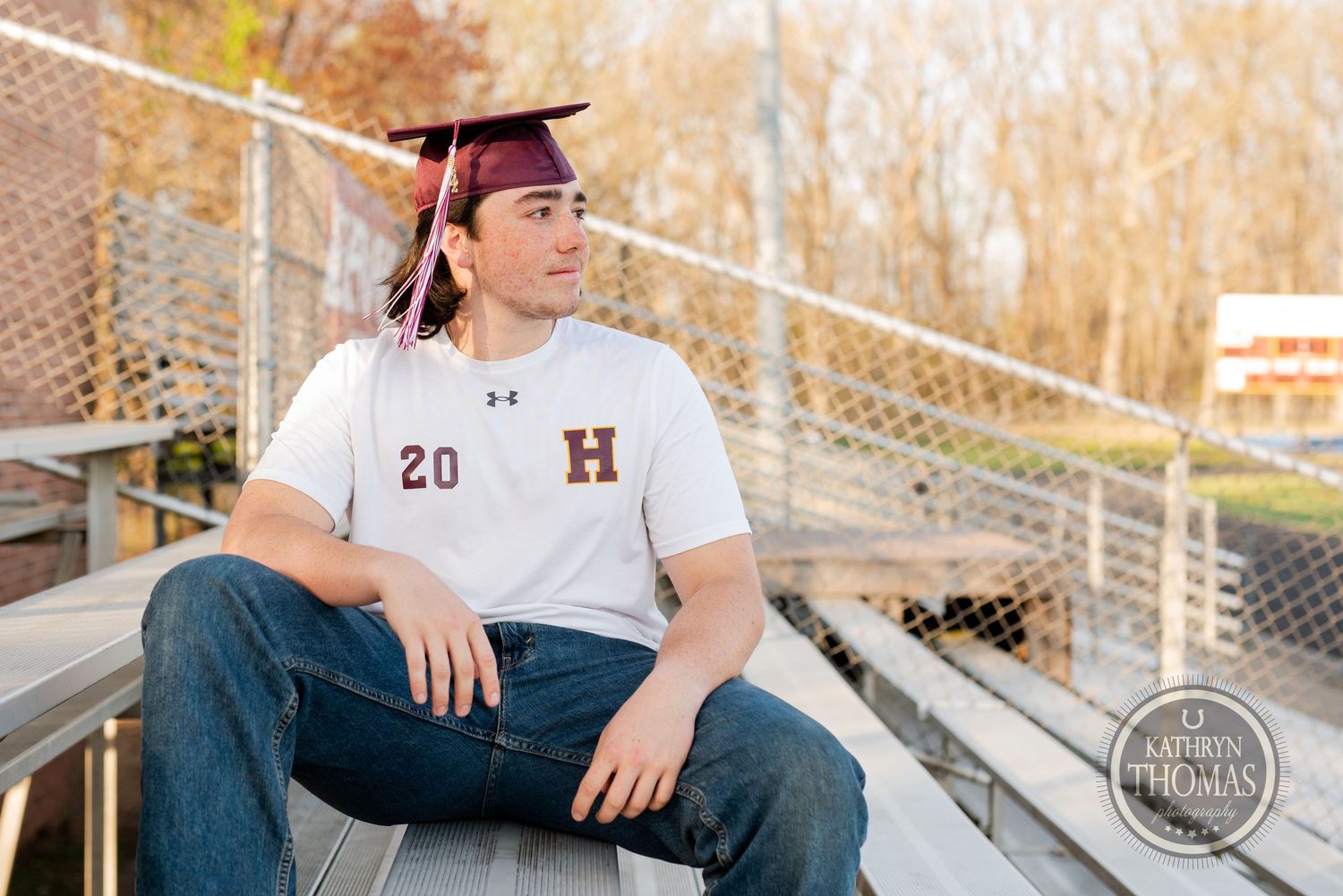 Graduate in cap and jersey relaxes on metal bleachers during outdoor senior photo session.
