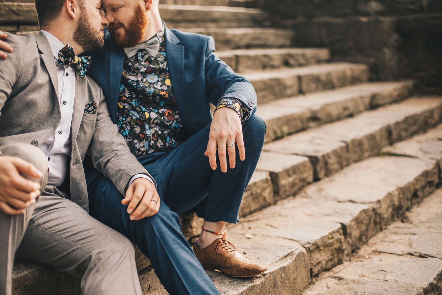 Two grooms in suits sit closely together on stone steps during their wedding.