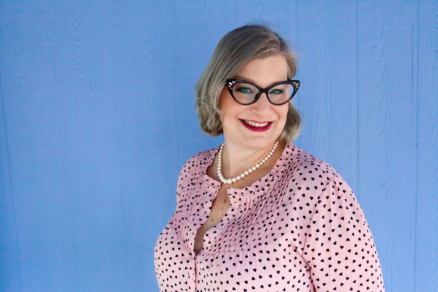 A blonde woman in a pink shirt from bust up, wearing black glasses against a purple wall