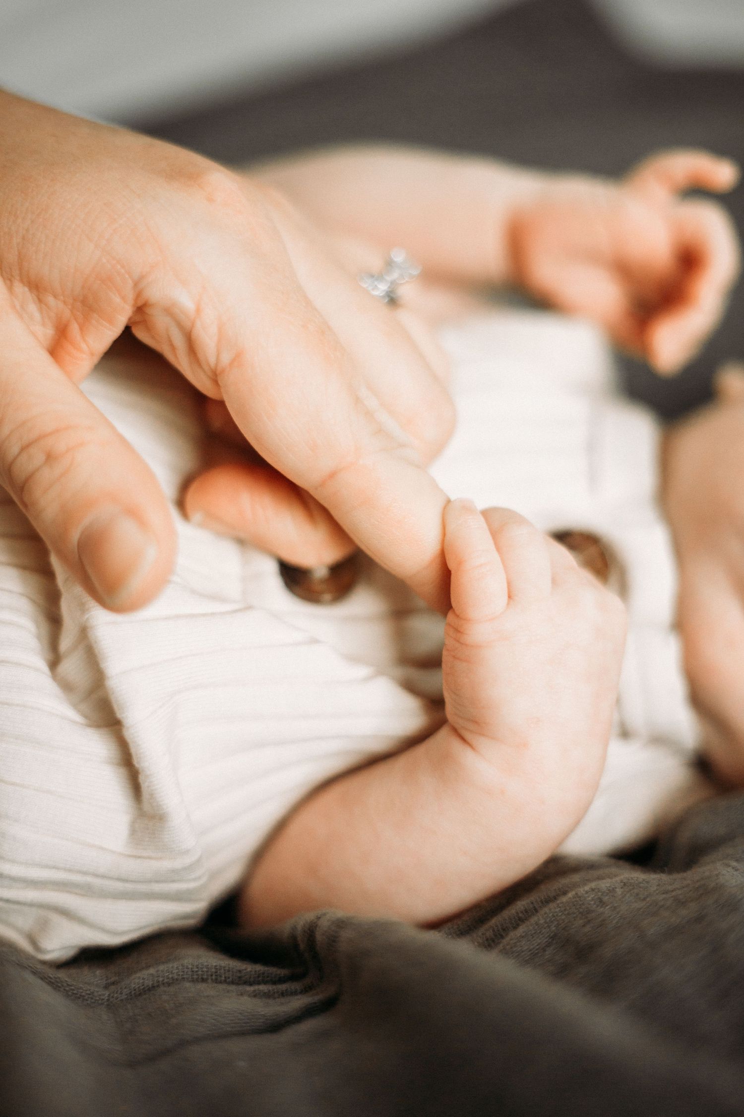 Close-up of a tiny baby hand grasping an adult's finger while lying on grey bedding.