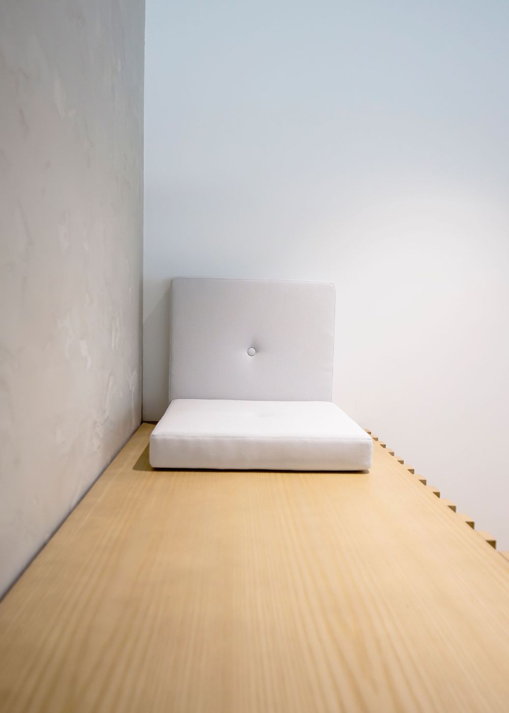 Minimalist white cushioned seat against a plain wall with wooden flooring.
