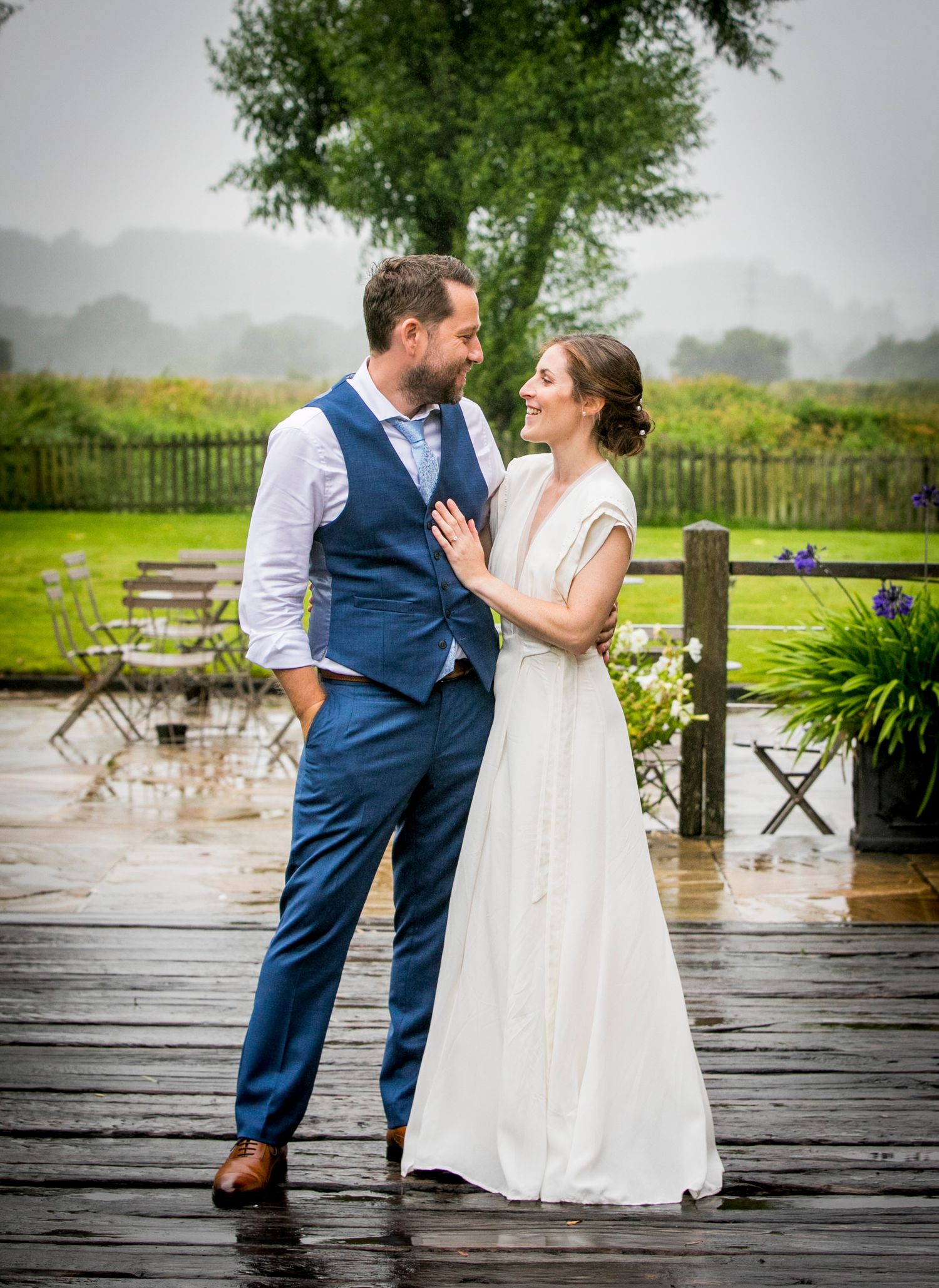 Couple in wedding attire shares intimate moments on a wet wooden deck after rain.