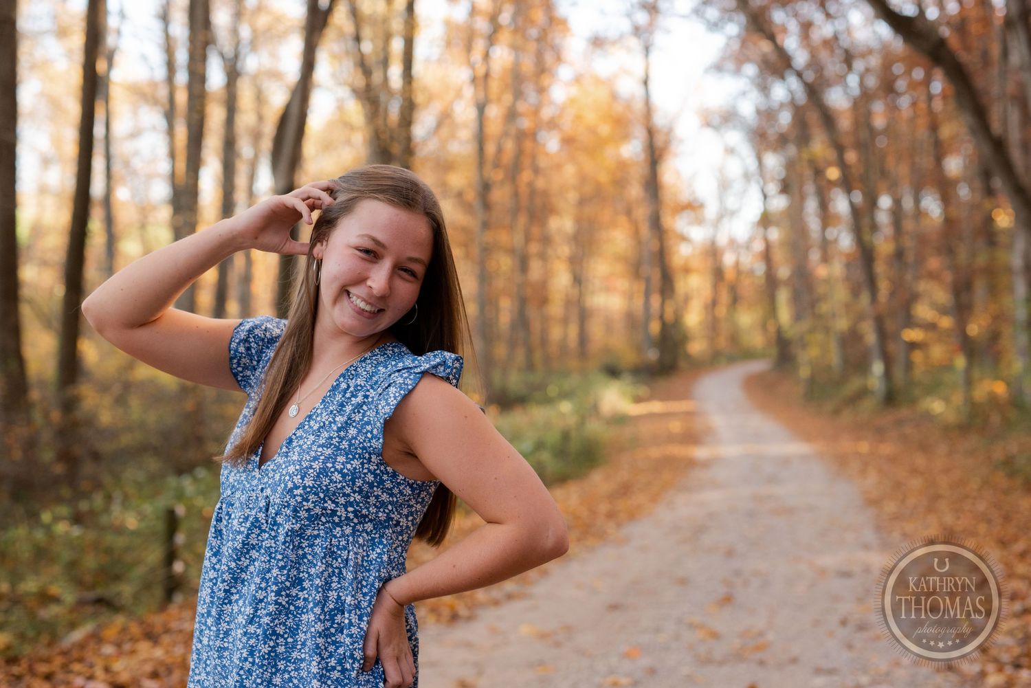 A person in blue dress poses playfully on a forest trail surrounded by fall colors.