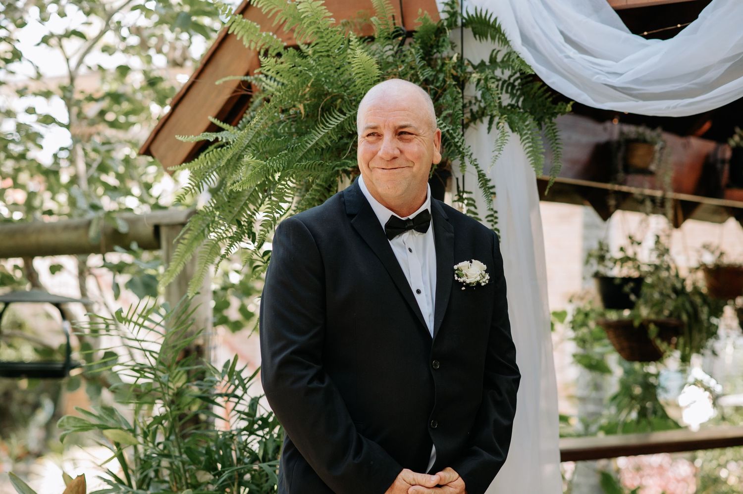 Groom in black tuxedo stands under decorated wedding arch.