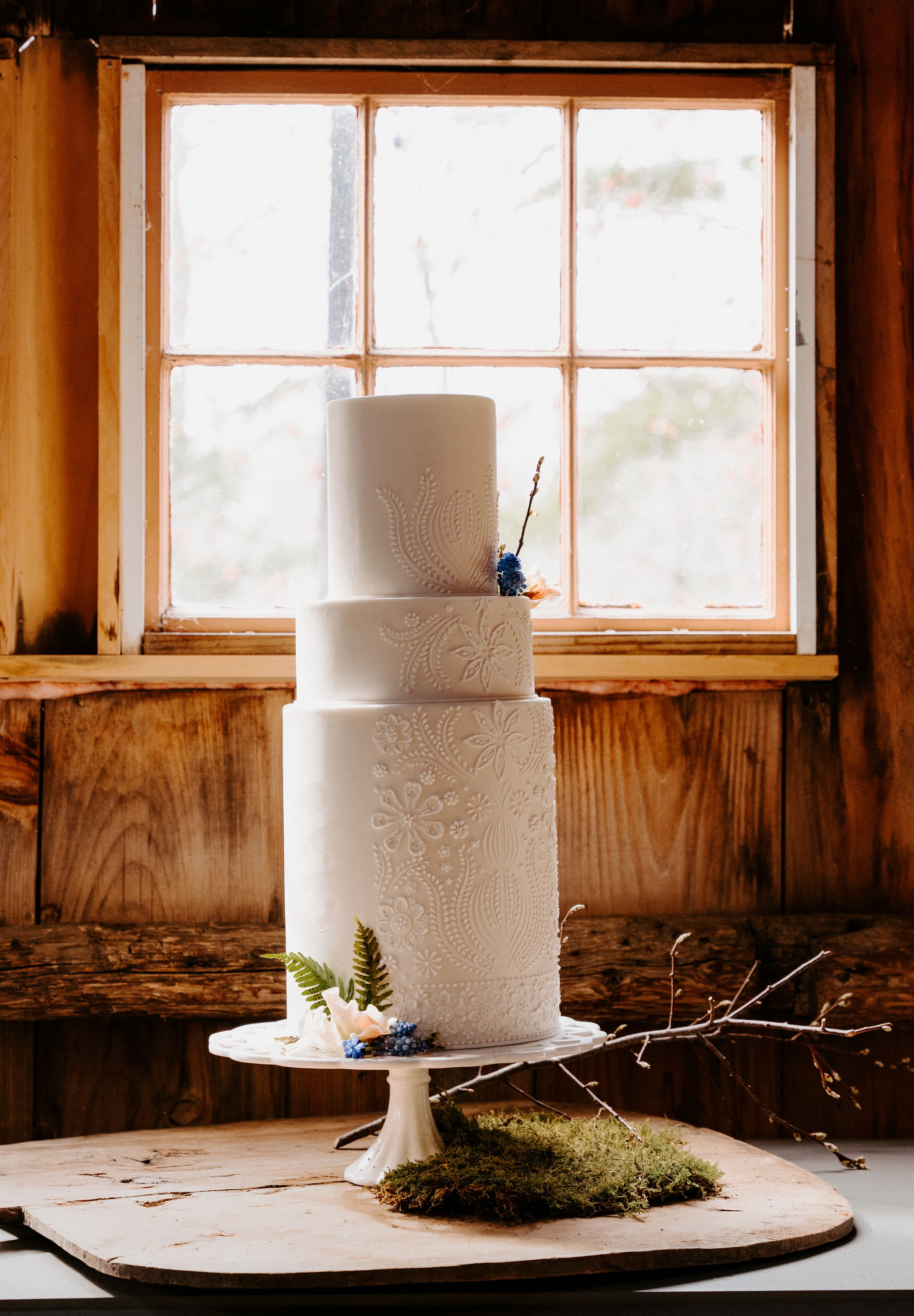 An elegant three-tiered white wedding cake decorated with delicate patterns sits near a window in rustic setting.