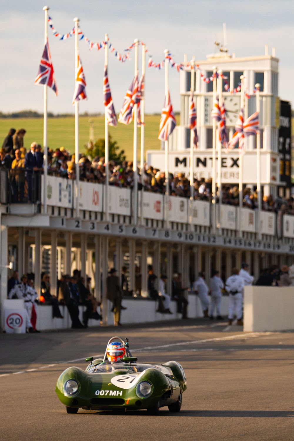 Classic racing car speeds past grandstands decorated with British flags during a vintage motorsport event.